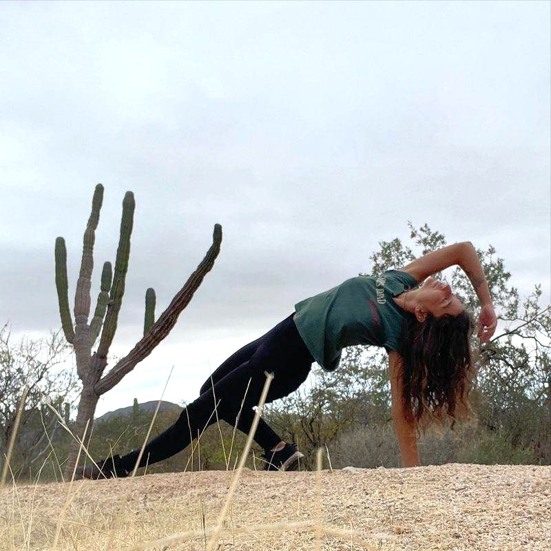 Shahad in a sideways stretch on a desert trail, beside a tall green cactus under a cloudy sky