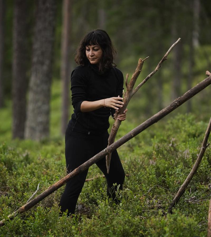 Sumaya in a green pine forest, carrying a long branch with both hands